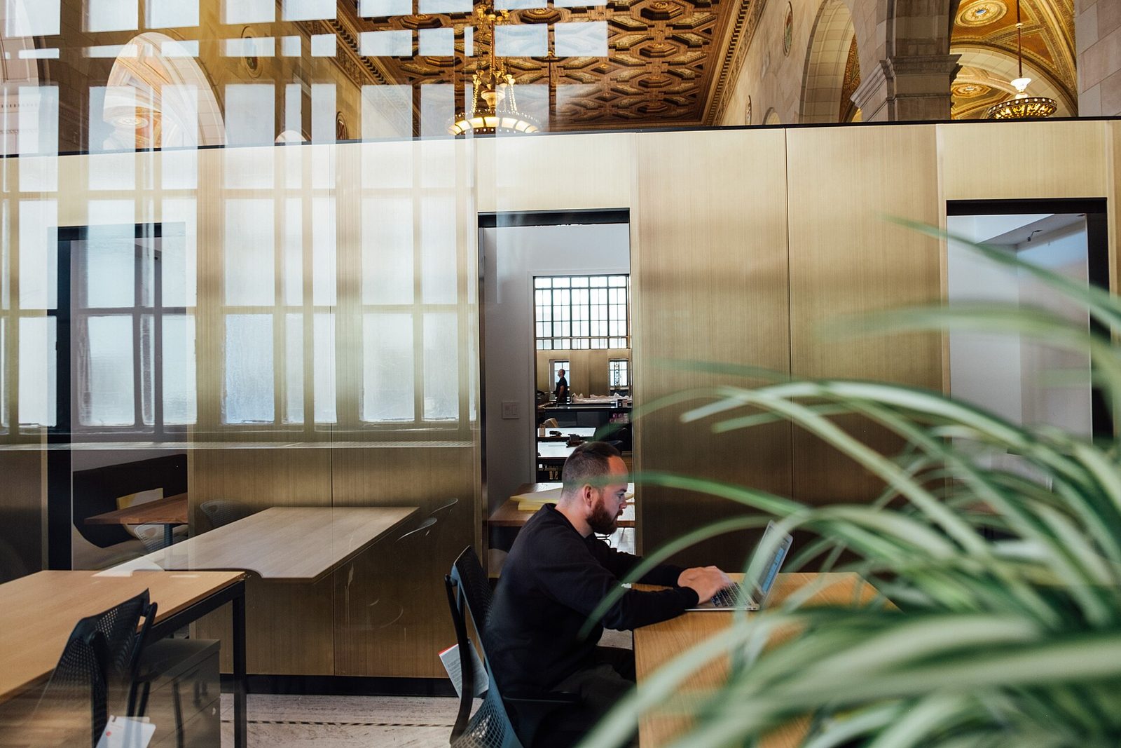 Personne travaillant sur un ordinateur portable dans un espace de bureaux aux cloisons vitrées et dorées, avec des plantes au premier plan