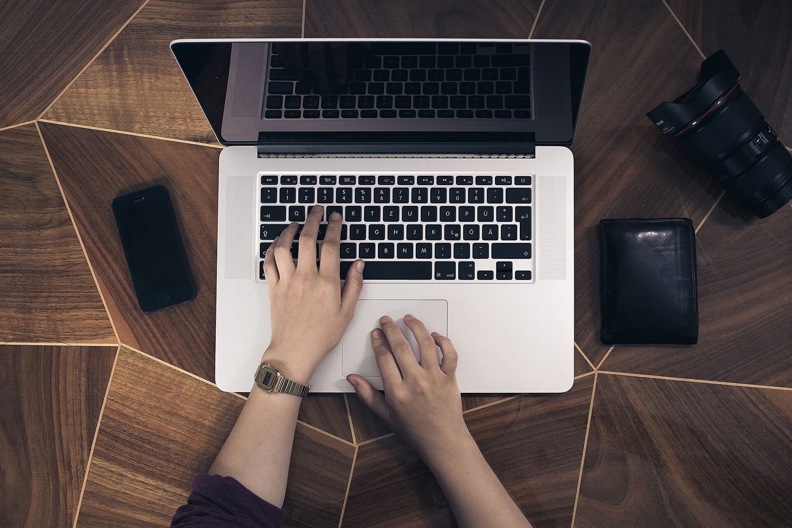 Mains sur le clavier d’un ordinateur portable posé sur un bureau en bois, avec un smartphone et un appareil photo