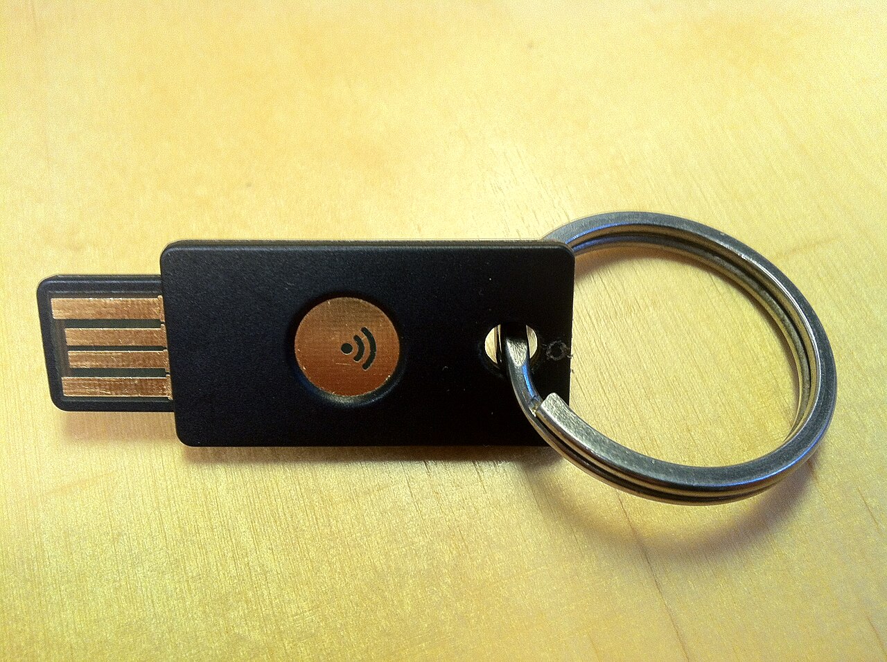 Clé de sécurité USB noire de type YubiKey, avec bouton tactile et anneau porte-clés, posée sur un bureau en bois