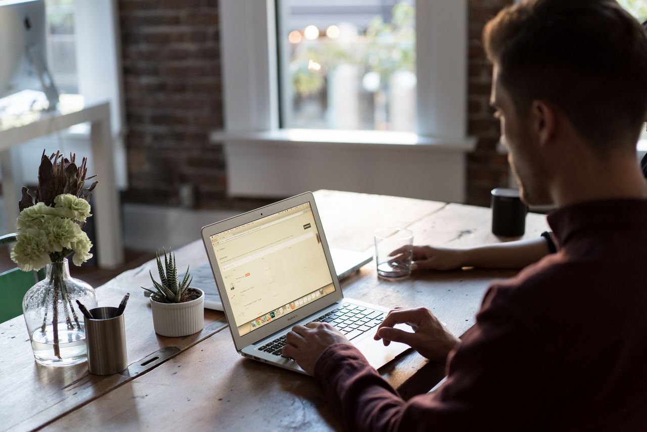 Homme travaillant sur un ordinateur portable posé sur une table en bois, dans un petit bureau aux murs de briques, avec plantes et verre d’eau